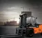 Front angled view of orange and black Doosan forklift with extended forks and enclosed cabin outdoors on a construction site.