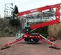 Side view of a red Teupen tracked atrium lift with basket positioned on brick pavement in an outdoor work zone.