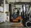 Side view of orange and grey Toyota forklifts with in a warehouse, one has an operator seated and one is in partial view.