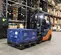 Front angled view of an orange and black warehouse forklift with an operator in the cabin, lifting a crate in a warehouse.