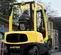 A man operates a yellow and black Hyster 50 forklift, loading a caged pallet with red cylinders onto a dark grey loading dock or ramp. The forklift is viewed from the rear-right side, with a person in the driver's seat, and trees and buildings in the background.